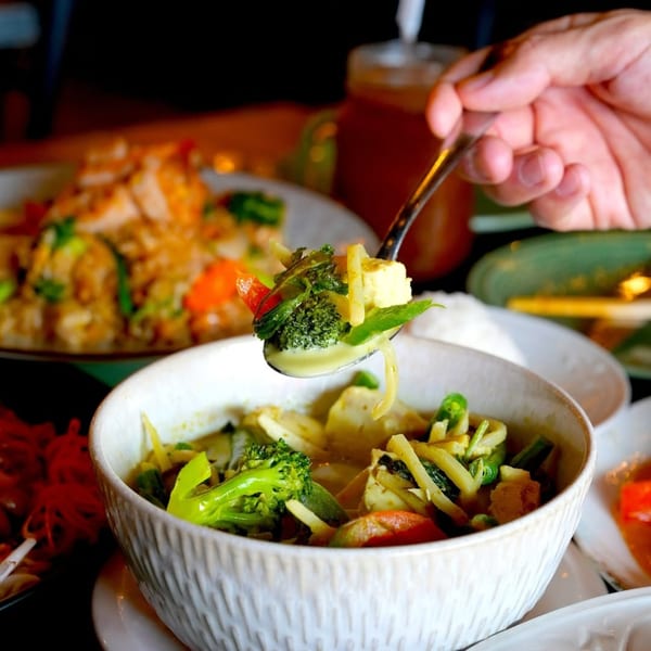 A hand spoons green curry with broccoli, bamboo shoots, and tofu from a white bowl at 8thai.