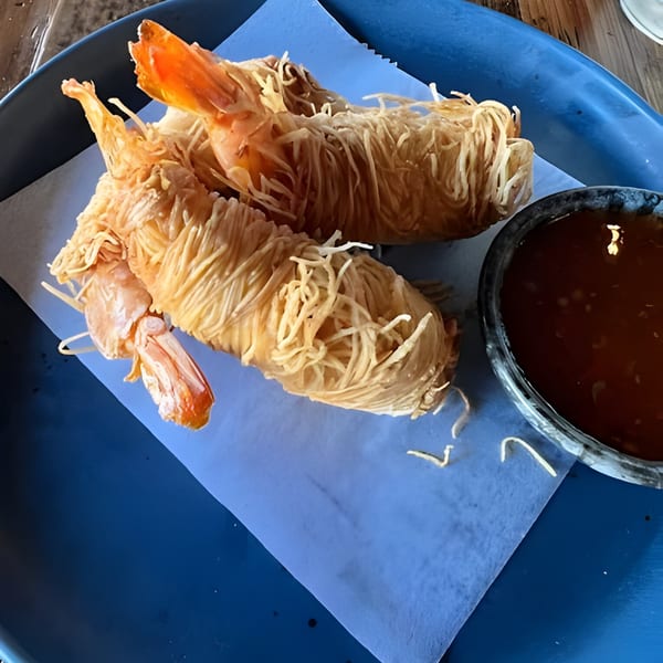 Two golden-fried shrimp wrapped in crispy noodles, served with a dark dipping sauce on a blue plate.