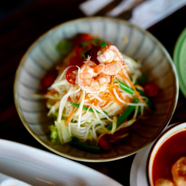 A bowl of Thai papaya salad with shrimp, noodles, tomatoes, and green beans.