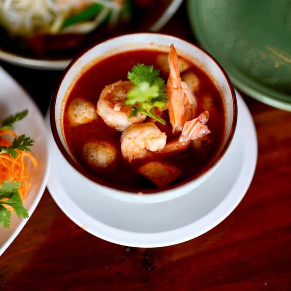 A bowl of vibrant red Tom Yum soup with shrimp, fish balls, and cilantro garnish.
